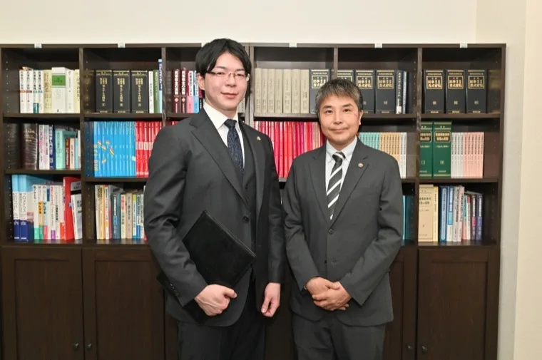 法律書が並ぶ執務室に立つ茨城県の交通事故に強いつくば第一法律事務所の弁護士2名